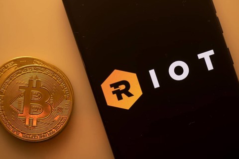 Bitcoin coin next to phone showing Riot Blockchain logo with black and hexagonal design