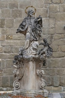 Baroque stone statue of a saint with angel on a decorated pedestal in front of a stone wall, parish