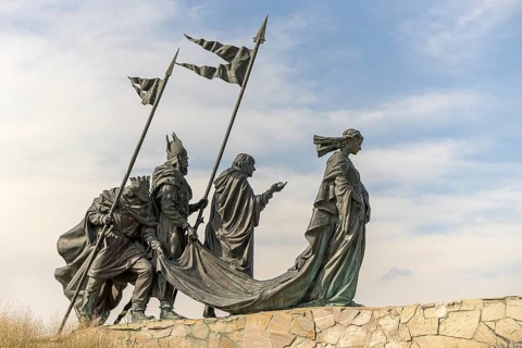 Sculpture group of historical figures with flags on a stone platform under a blue sky,