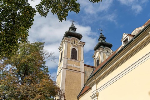 Baroque church towers with clocks in front of a blue sky, surrounded by autumnal trees, parish