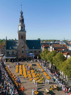 Market square with cheese wheels in front of a church and many visitors in sunny weather, aerial