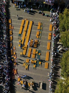 Aerial view of a busy market square with lots of cheese wheels and people at a sunny festival,