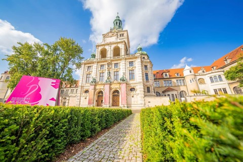 Historic building with towers and pink banners framed by manicured hedges and blue sky, Munich,