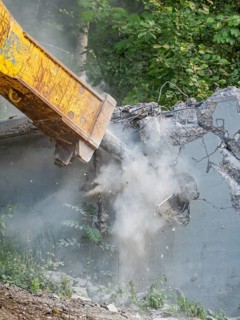 An excavator tears down a concrete structure with its shovel, surrounded by forest, dusty