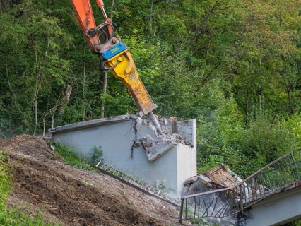 An excavator destroys a concrete wall on a slope, remains of a ladder visible, bridge demolition,