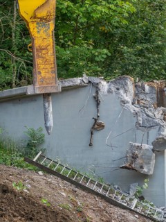 A demolition hammer destroys concrete, a ladder leans against the wall, surrounded by plants,