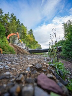 Wide angle shot of an excavator on a gravel road demolishing a bridge, surrounded by lush greenery