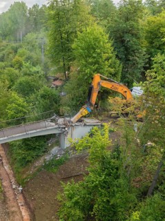 Excavator tears down part of a bridge in a green forest, an empty path runs underneath, bridge