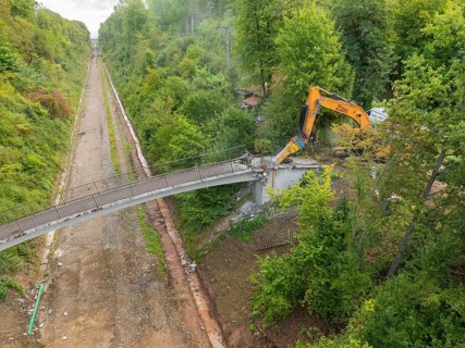 Bird's eye view of an excavator demolishing a bridge in the middle of the forest, with an empty