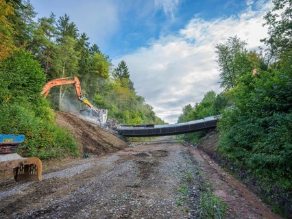 Two excavators tearing down a bridge on a gravel road, flanked by forest and a cloudy sky, bridge