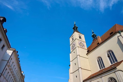 Gothic-style church tower and building under a bright blue sky, flanked by other buildings,