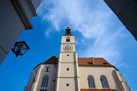 View of a high church tower with a clock under a bright blue sky, flanked by buildings,