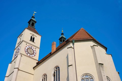 Close-up of a church tower with clock and red tiled roofs against a deep blue sky, Neupfarrkirche,