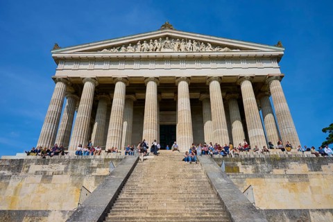 Temple-like structure with monumental columns and visitors on a staircase under a blue sky,