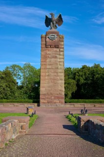 Monument with eagle on a pedestal, surrounded by trees. Wide paved path leads to the memorial.