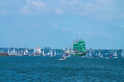 Green tall ship Alexander von Humbold 2 surrounded by many boats in front of an urban backdrop,