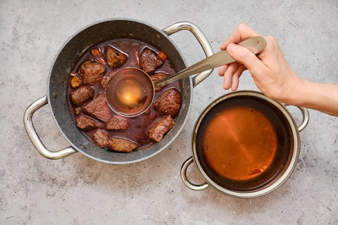 A person is using a ladle to add beef broth to a pot filled with fried chunks of beef shoulder and