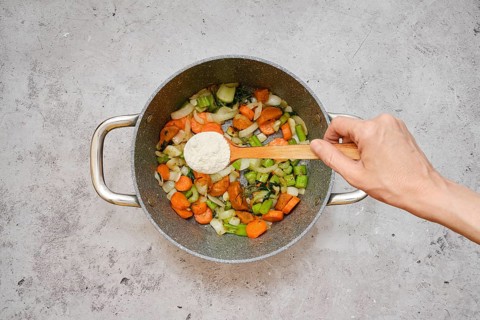 Freshly chopped carrots, celery, and onions are fried in a pot. A hand is sprinkling flour over the