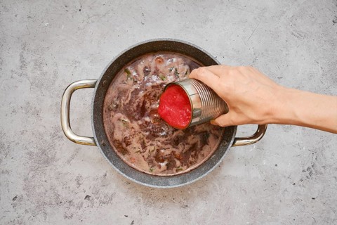 A hand pours diced tomatoes from a can into a pot filled with a rich, simmering beef stew