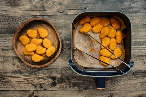 Golden and crispy chicken nuggets rest in an air fryer beside a wooden plate filled with freshly
