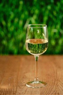 A clear glass filled with white wine sits on a smooth wooden table. The soft green background