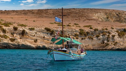 A boat with a Greek flag sails along rocky shores, the sea is blue and calm, White Cliffs, White