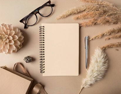 Flat lay composition featuring a beige spiral notebook, elegant pen, glasses, dried flowers, and