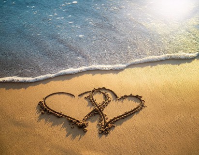 Warm tropical beach with two hearts drawn on the sand, blue waters, and clear skies, invoking