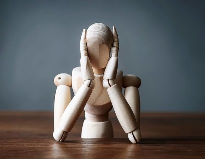 A wooden mannequin seated at a table conveys emotions of stress, thought, and contemplation.