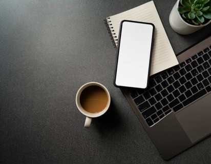 Op view of an organized workspace featuring a laptop, smartphone, notebook, pen, plant, and coffee