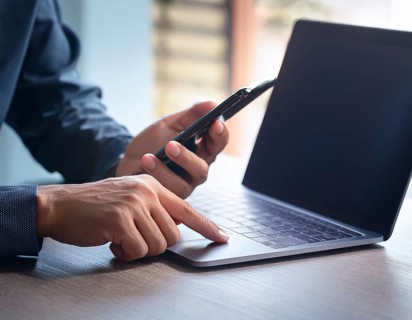 A man in a professional office environment multitasking with a smartphone and laptop, conveying