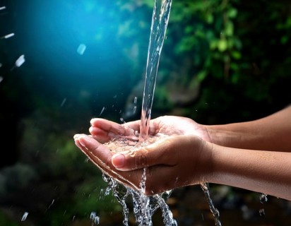 A pair of human hands collecting fresh water flowing from a source, surrounded by grassy landscape,