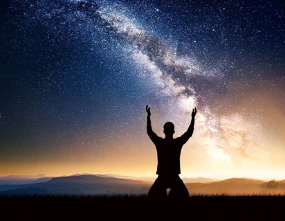 A man kneels in peaceful reflection under the vibrant Milky Way galaxy at sunset in an open natural