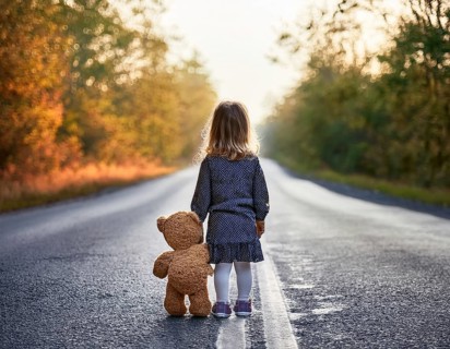 A young child stands alone holding a teddy bear in an urban outdoor setting, symbolizing innocence