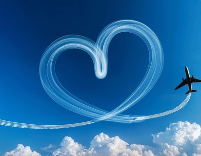An airplane creates heart-shaped contrails in a vivid blue sky above fluffy white clouds,