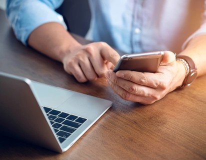 A man in a professional office environment multitasking with a smartphone and laptop, conveying