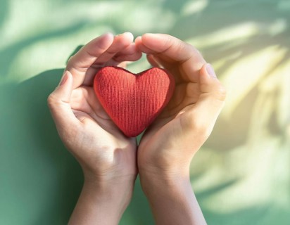 A pair of hands gently holding a red fabric heart, conveying themes of love, care, and compassion.