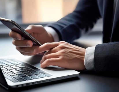 A man in a professional office environment multitasking with a smartphone and laptop, conveying