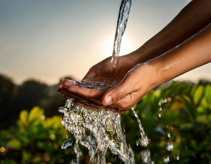 A pair of human hands collecting fresh water flowing from a source, surrounded by grassy landscape,