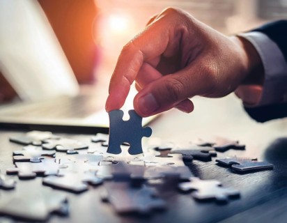 A person's hand placing a puzzle piece on a reflective surface in an office setting as the sun