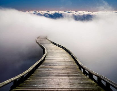Curved wooden boardwalk disappearing into a thick white mist under a calm sky. This scenic view