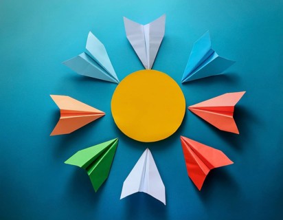 Six colorful origami paper airplanes are displayed in a creative arrangement on a bright