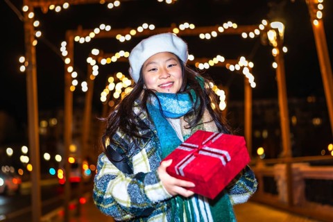 Smiling young woman holding christmas present enjoying christmas lights decoration at night
