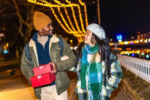Young multiethnic couple walking arm in arm at illuminated christmas market, man carrying christmas