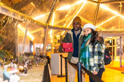 Multiethnic couple holding christmas gifts looking at a nativity scene in a christmas village
