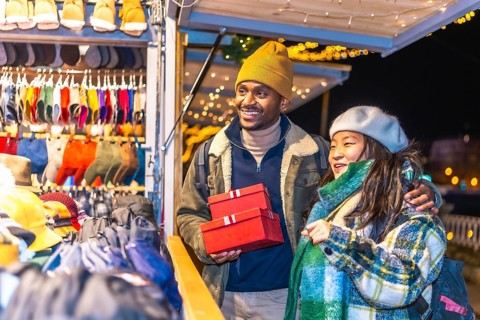 Smiling multiethnic couple holding christmas gifts while enjoying christmas market at night