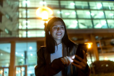 Young woman smiling and engaging with her smartphone, wearing wireless earbuds for connectivity and