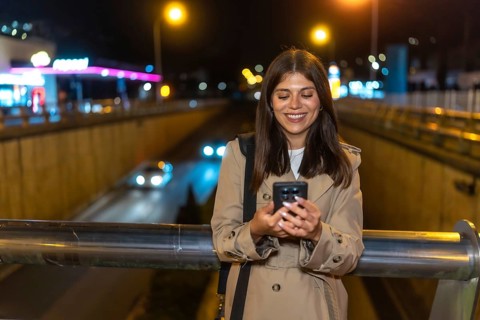 Young woman smiling while looking at her smartphone, standing on an urban bridge with city lights