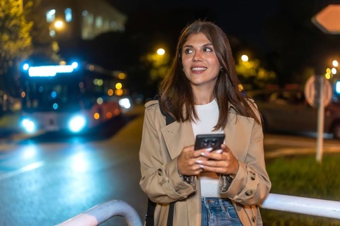 Young woman smiling while checking her mobile phone at a city bus stop at night, anticipating