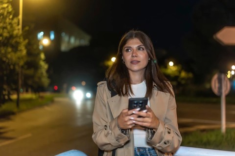 Young woman standing on city street at night, holding smartphone and looking away as warm car